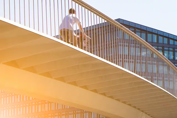 A man riding a bicycle on a footbridge wth a modern building and greenery in the background