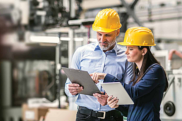 Two people in safety equipment looking at a clipboard with a blurred factory background