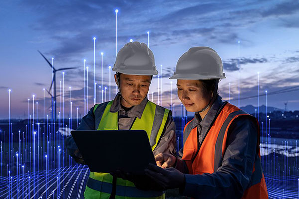 Two people in safety equipment looking at a laptop with wind turbines in the background